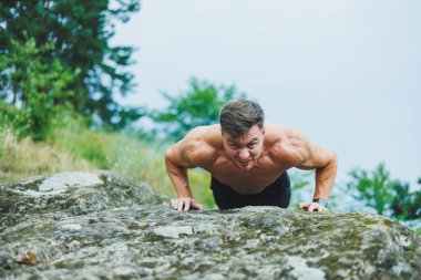Ormanın arka planında karın kasları olan genç bir adam. Kayalık zeminde atletik şişmiş bir adam.