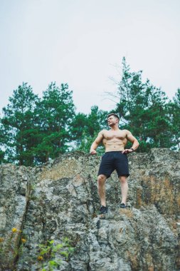Ormanın arka planında karın kasları olan genç bir adam. Kayalık zeminde atletik şişmiş bir adam.