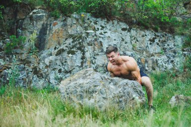 Bir sporcunun fotoğrafı, kaslı, atletik bir genç adam çıplak bir gövdeyle egzersiz yapıyor, ormanda, dağlarda..