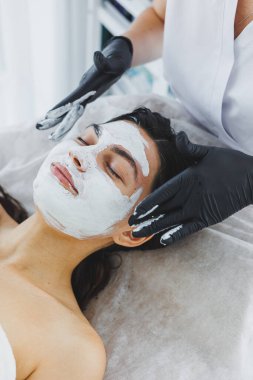 Bir kadının yüzü için cilt bakımı. Yüzünde beyaz maske olan bir kadın güzellik uzmanının kanepesinde yatıyor. Güzellik salonunda kozmetoloji ve yüz bakımı. Beyaz maske.