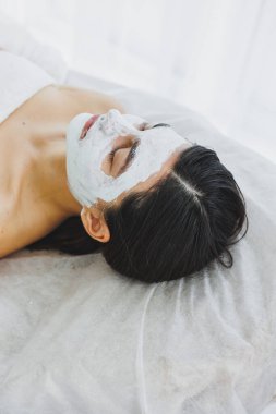 Bir kadının yüzü için cilt bakımı. Yüzünde beyaz maske olan bir kadın güzellik uzmanının kanepesinde yatıyor. Güzellik salonunda kozmetoloji ve yüz bakımı. Beyaz maske.