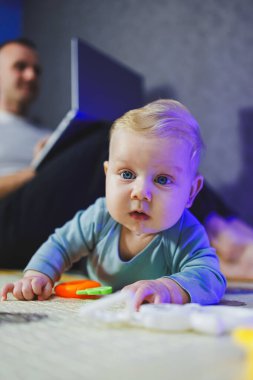 Küçük oğlu yanında oynarken endişeli bir baba evde dizüstü bilgisayarla çalışıyor. Yerde bir bebek oynuyor..