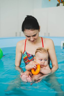 Bir bebek havuzda koçla yüzmeyi öğrenir. Küçük çocuklar için yüzme kursları. Çocuklar için yüzme havuzu