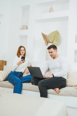 A man and a woman are sitting on the couch at home and watching a video on a laptop.