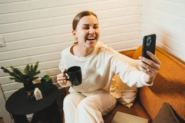 stock image A woman in home clothes is sitting at home on the sofa with a laptop and talking on the phone. Work from home.