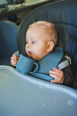 Bebek yüksek bir sandalyede otururken silikon bardaktan su içer. Bebekler için sofra takımı.