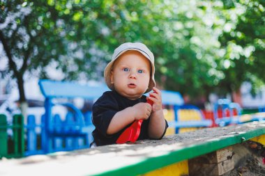 Kafasında panama olan bir erkek bebek oyun parkında oynuyor. Sokakta yürüyen bir çocuk.
