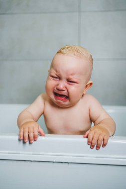 Erkek bebek, beyaz bir küvette suyla birlikte evde banyo yapıyor. Bebek gülümsüyor ve mutlu su sıçrıyor..