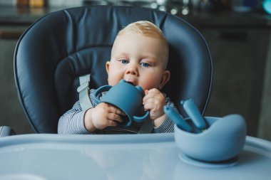 Bebek bir sandalyede otururken silikon bardaktan su içer. Bebekler için sofra takımı.