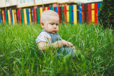 Bir yaşında küçük bir çocuk sokakta yeşil çimlerde oturuyor. Bebekler için yaz giysileri.