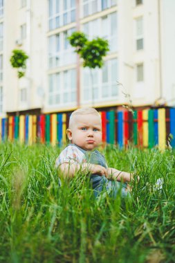Bir yaşında küçük bir çocuk sokakta yeşil çimlerde oturuyor. Bebekler için yaz giysileri.