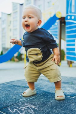Bir yaşındaki küçük bir çocuk sokakta tebeşirle resim çiziyor. Bebekler için yaz giysileri. Sandaletli bir çocuk..