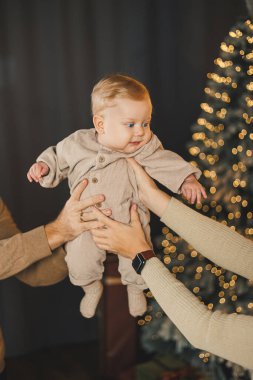 Noel kıyafetli küçük bir çocuk bir Noel ağacının arka planında oturuyor. Evdeki Noel ağacının yanındaki küçük çocuk..