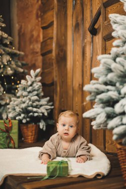 Noel ağacının arkasında paketlenmiş bir hediyeyle bir bebek. Yılbaşı süslemelerinin arka planında evde bir çocuk.