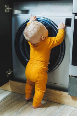 Mavi gözlü, açık renk elbiseli küçük bir çocuk çamaşır makinesine bakıyor..