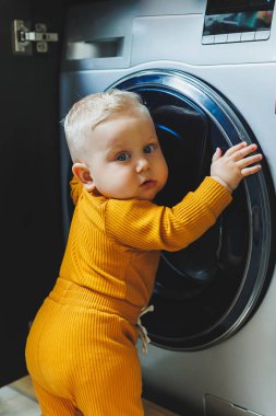 Mavi gözlü, açık renk elbiseli küçük bir çocuk çamaşır makinesine bakıyor..