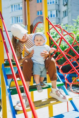 Kafasında tişört ve panama olan 11 aylık bir oğlan çocuğu annesiyle oyun parkında oynuyor. Bir çocuk ve annesi sokakta oynuyorlar. Bebekler için yaz giysileri