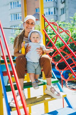 Kafasında tişört ve panama olan 11 aylık bir oğlan çocuğu annesiyle oyun parkında oynuyor. Bir çocuk ve annesi sokakta oynuyorlar. Bebekler için yaz giysileri
