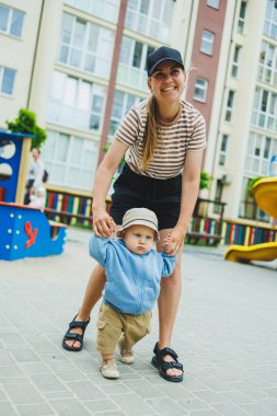 Bir anne oğluna yürümeyi öğretir, bir bebeğin ilk adımları. Çocuk annesinin elini tutarak yürür. Kapüşonlu bir bebek ve kısa bir yürüyüş. Erkek çocuklar için kıyafetler..