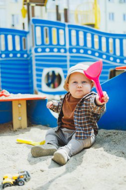 Küçük sevimli bir çocuk kum havuzunda otururken bir kürekle oynuyor. Bir çocuk kumla oynar. Sokakta yürüyen bir bebek. Çocuğun kişisel gelişimi. Bir erkek için çocuk kıyafetleri..