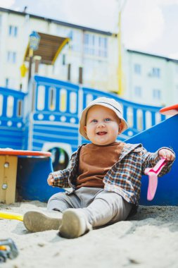 Küçük sevimli bir çocuk kum havuzunda otururken bir kürekle oynuyor. Bir çocuk kumla oynar. Sokakta yürüyen bir bebek. Çocuğun kişisel gelişimi. Bir erkek için çocuk kıyafetleri..
