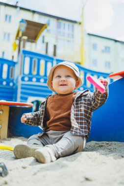 Küçük sevimli bir çocuk kum havuzunda otururken bir kürekle oynuyor. Bir çocuk kumla oynar. Sokakta yürüyen bir bebek. Çocuğun kişisel gelişimi. Bir erkek için çocuk kıyafetleri..