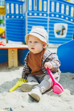 Küçük sevimli bir çocuk kum havuzunda otururken bir kürekle oynuyor. Bir çocuk kumla oynar. Sokakta yürüyen bir bebek. Çocuğun kişisel gelişimi. Bir erkek için çocuk kıyafetleri..
