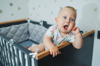 Mavi gözlü tişörtlü bir bebek beşiğindeki korkulukların yanında duruyor. Bebeğin hisleri işte. Cot bir çocuk için..