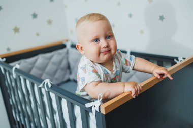 Mavi gözlü tişörtlü bir bebek beşiğindeki korkulukların yanında duruyor. Bebeğin hisleri işte. Cot bir çocuk için..