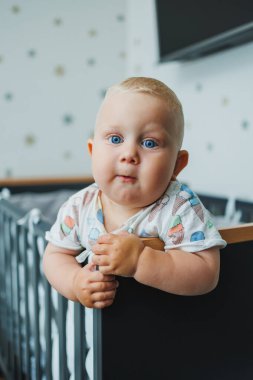 Mavi gözlü tişörtlü bir bebek beşiğindeki korkulukların yanında duruyor. Bebeğin hisleri işte. Cot bir çocuk için..