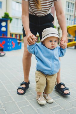 Bir anne oğluna yürümeyi öğretir, bir bebeğin ilk adımları. Çocuk annesinin elini tutarak yürür. Kapüşonlu bir bebek ve kısa bir yürüyüş. Erkek çocuklar için kıyafetler..