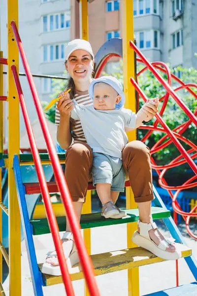 Kafasında tişört ve panama olan 11 aylık bir oğlan çocuğu annesiyle oyun parkında oynuyor. Bir çocuk ve annesi sokakta oynuyorlar. Bebekler için yaz giysileri