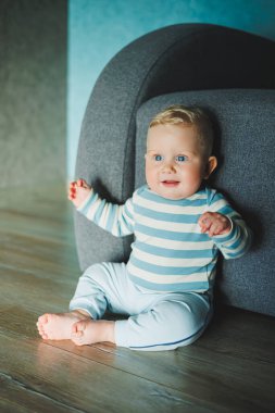 Yüzünde gülümseme olan mavi gözlü bir bebek. Eşofman ve pantolon giymiş bir bebek evde yerde oturuyor..