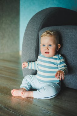 Yüzünde gülümseme olan mavi gözlü bir bebek. Eşofman ve pantolon giymiş bir bebek evde yerde oturuyor..
