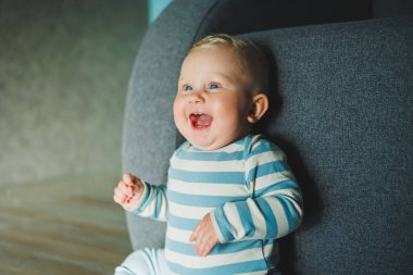 Yüzünde gülümseme olan mavi gözlü bir bebek. Eşofman ve pantolon giymiş bir bebek evde yerde oturuyor..