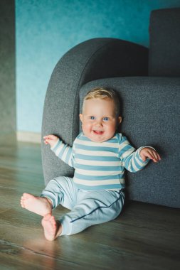 Yüzünde gülümseme olan mavi gözlü bir bebek. Eşofman ve pantolon giymiş bir bebek evde yerde oturuyor..