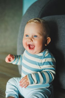 Yüzünde gülümseme olan mavi gözlü bir bebek. Eşofman ve pantolon giymiş bir bebek evde yerde oturuyor..