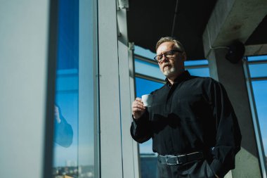 Yaşlı iş adamı elinde kahveyle pencerenin önünde duruyor. O, siyah bir gömlek ve gözlük takıyor. Bir işadamı gökdelendeki ofis penceresinden şehre bakıyor.