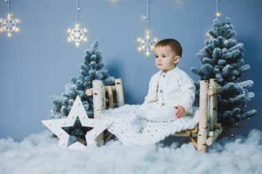 Beyaz örgü takım elbiseli küçük bir çocuk Noel ağacının yanında oturuyor. Yeni yıl havası. Çocuklar için örgü kostüm..