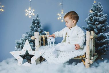 Beyaz örgü takım elbiseli küçük bir çocuk Noel ağacının yanında oturuyor. Yeni yıl havası. Çocuklar için örgü kostüm..