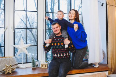 Sıcak örülü süveter giyen bir aile Noel süslemeleriyle pencere eşiğinde oturuyor. Samimi bir evde yeni yıl süslemeleri. Yeni yıl için aile pencerenin yanında.