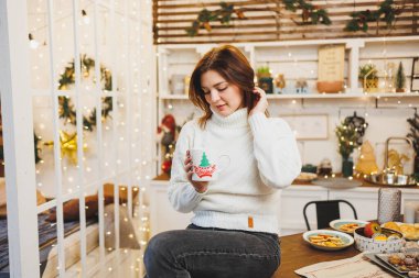 Beyaz örgü süveter giyen tatlı bir kadın sıcak bir mutfakta kahveyle Noel süslemeleri ile oturuyor. Yeni yıl tarzında mutfak..