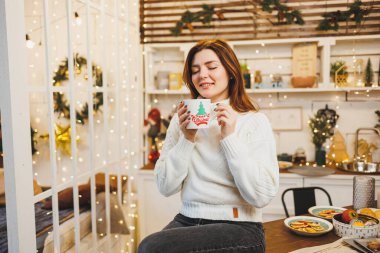 Beyaz örgü süveter giyen tatlı bir kadın sıcak bir mutfakta kahveyle Noel süslemeleri ile oturuyor. Yeni yıl tarzında mutfak..