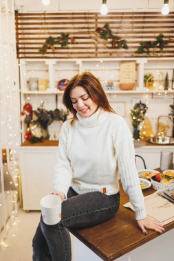 Beyaz örgü süveter giyen tatlı bir kadın sıcak bir mutfakta kahveyle Noel süslemeleri ile oturuyor. Yeni yıl tarzında mutfak..