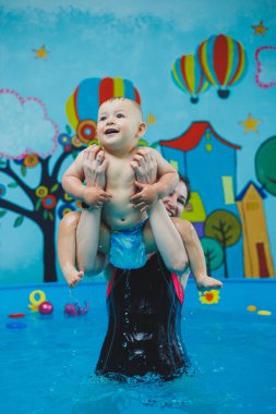 Bir antrenör küçük bir çocuğa çocuk havuzunda yüzmeyi öğretir. Bebeklere yüzmeyi öğretme kursları.
