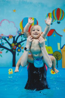 Bir antrenör küçük bir çocuğa çocuk havuzunda yüzmeyi öğretir. Bebeklere yüzmeyi öğretme kursları.