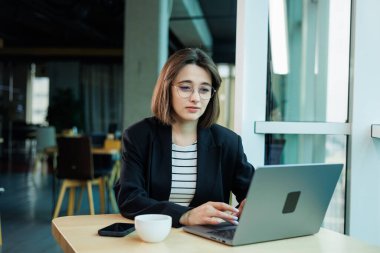 Gözlük takan genç bir kadın modern, ferah bir çalışma ofisinde oturuyor. Bilgisayarlı kadın ofis çalışanı çalışıyor..