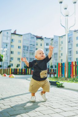 Çocuk parkında açık havada yaz elbisesi giymiş bir çocuk. Bir yaşında bir çocuk, yüksek binaların yakınındaki modern bir avluda yürüyor. Çok katlı bir binada çocuk parkı.
