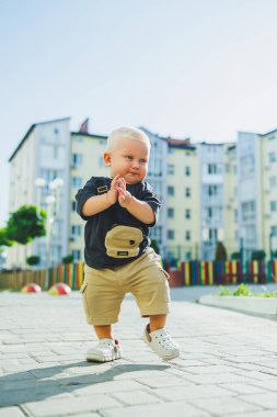 Çocuk parkında açık havada yaz elbisesi giymiş bir çocuk. Bir yaşında bir çocuk, yüksek binaların yakınındaki modern bir avluda yürüyor. Çok katlı bir binada çocuk parkı.