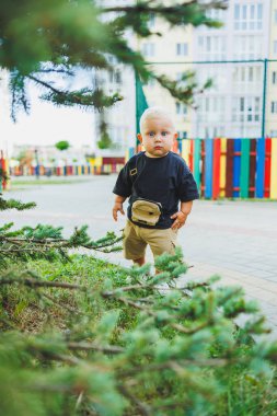 Çocuk parkında açık havada yaz elbisesi giymiş bir çocuk. Bir yaşında bir çocuk, yüksek binaların yakınındaki modern bir avluda yürüyor. Çok katlı bir binada çocuk parkı.